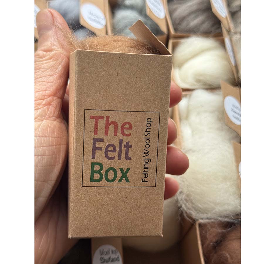 Small box with individual wool sample. Hand holding a small brown box labeled 'The Felt Box' with felt wool in the background.