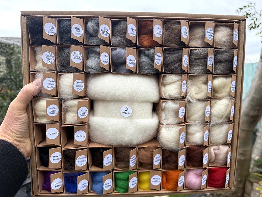 Box of assorted wool tops with labels held by a hand outdoors.