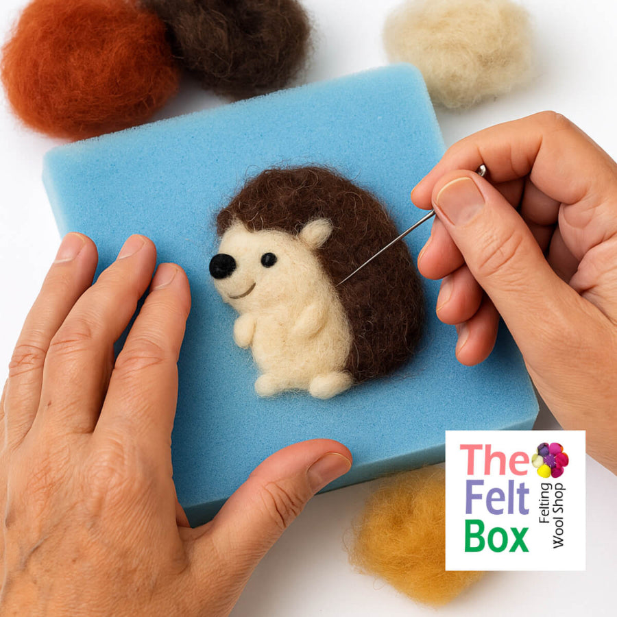 Person needle felting a hedgehog on a blue foam block with colored felts around, featuring 'The Felt Box' logo.